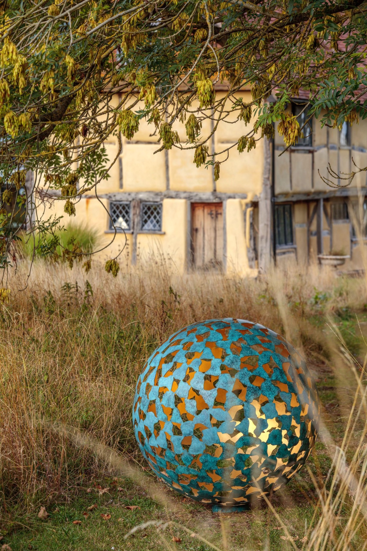 A pierced sphere by David Harber in long grass before a timbered house