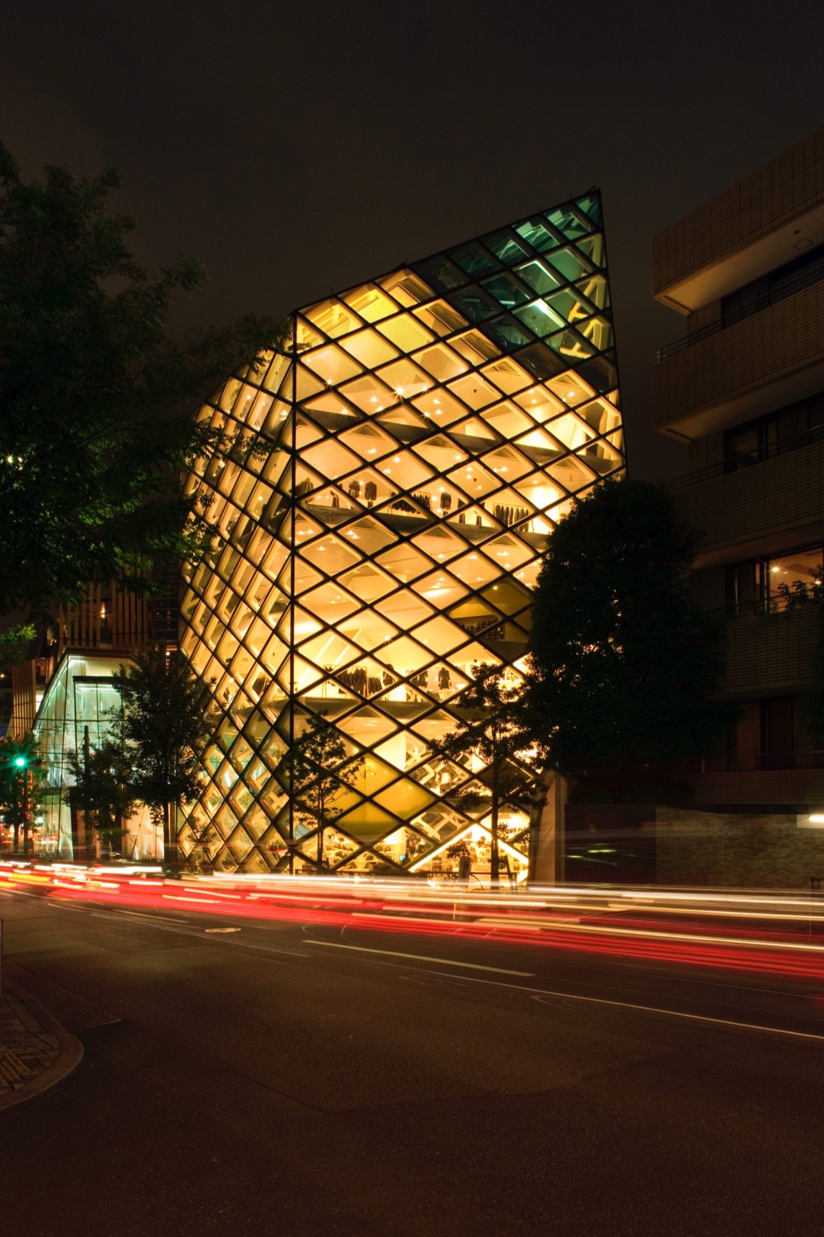 The Prada store in Aoyama, Tokyo, its diamond glass lit from within, traffic drawing light trails past it